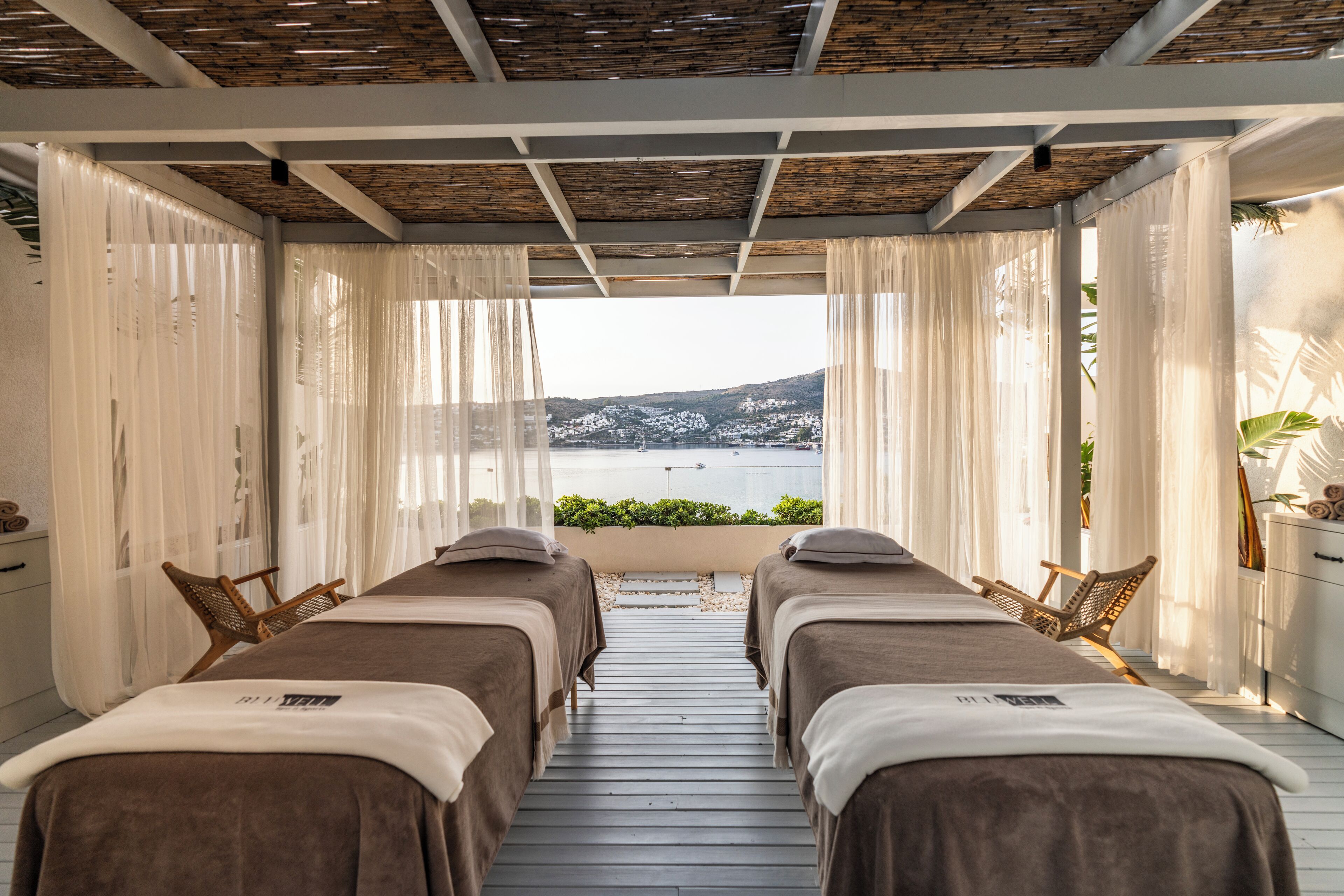 A spa room with two massage beds, a wooden ceiling, and a view of a body of water and hills.