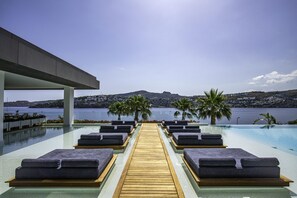 Zona junto a la piscina con sillones de descanso y una terraza de madera que da al agua, con vistas a un paisaje pintoresco con colinas y una masa de agua.