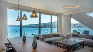 Een moderne woonkamer met een houten eettafel, banken en uitzicht op zee met boten.