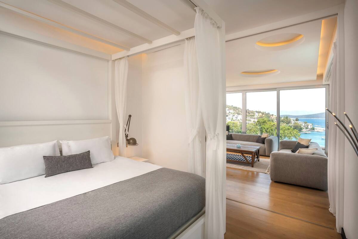 A modern bedroom with a large bed, a balcony with a view of the sea, and a seating area with a table.