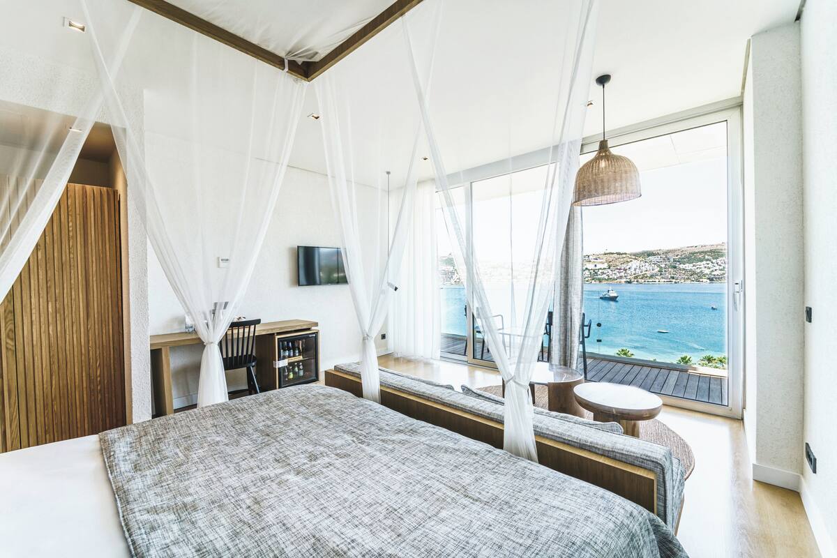 A bedroom with a large bed, a TV, a small table, and a view of the sea.