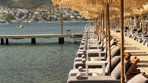 A seaside resort with rows of sun loungers and thatched umbrellas, a pier extending into calm waters, and a backdrop of a coastal town. - Cape Bodrum Luxury Hotel & Beach (Bodrum)