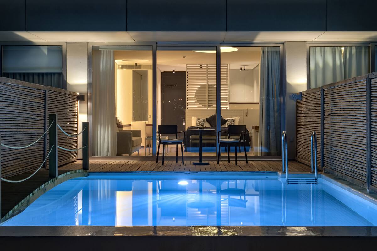 A modern indoor pool area with a wooden deck, a seating area with chairs and a table, and a bedroom visible through a glass door.