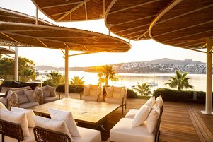 An outdoor seating area with a wooden table and wicker chairs, overlooking a body of water and a cityscape.