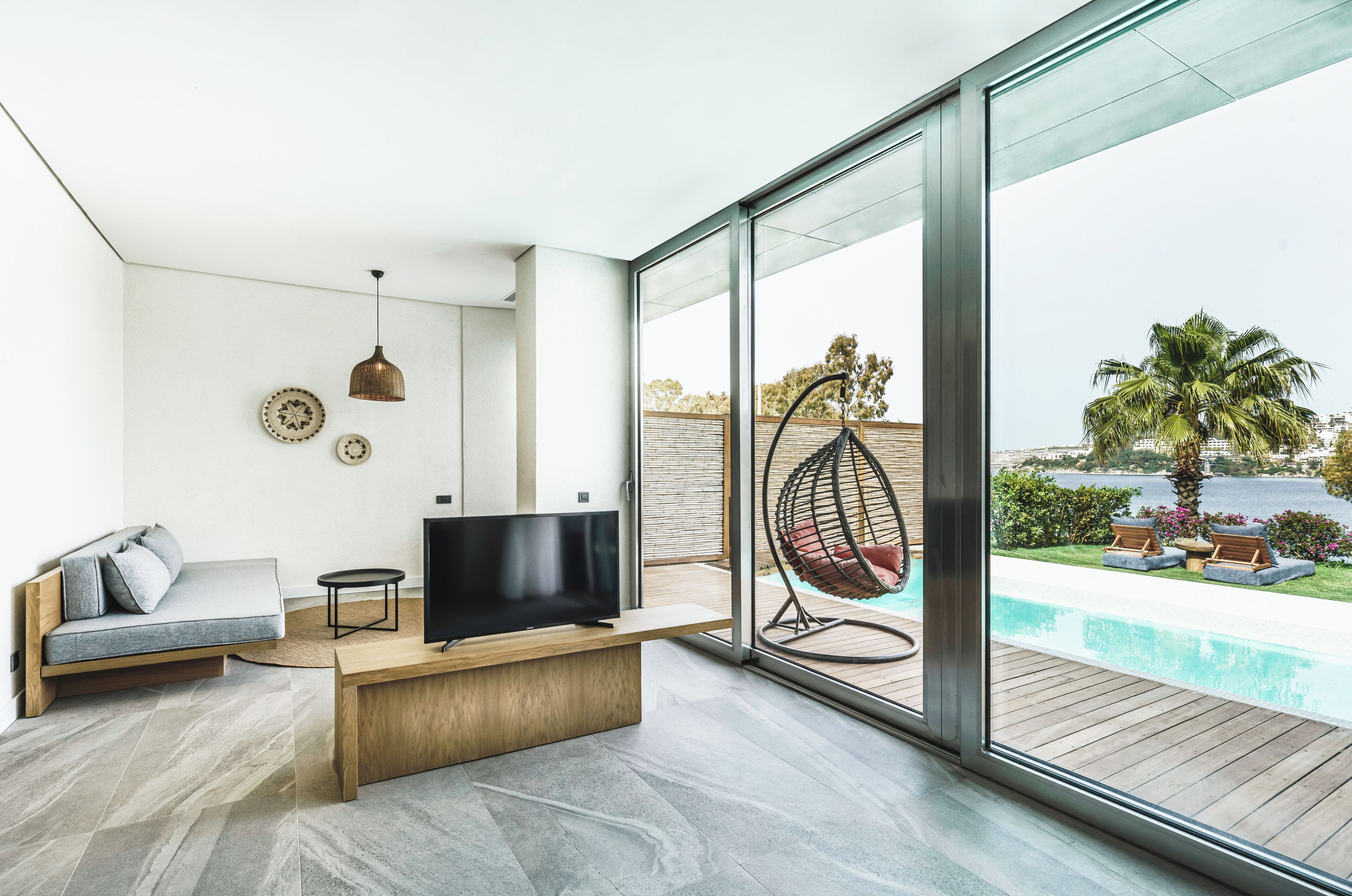 A modern living room with a TV, sofa, and hanging chair by a glass wall overlooking a pool.