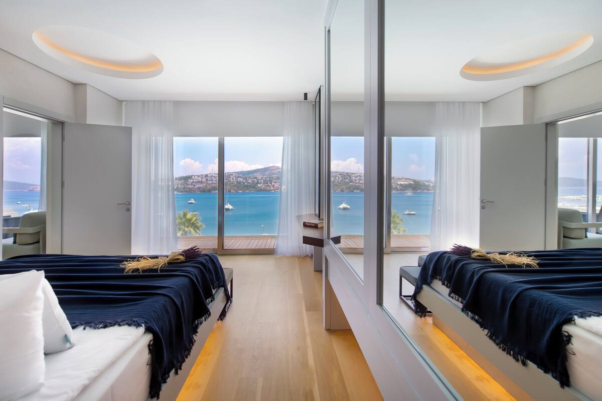 A hotel room with two beds, a large window with a view of the sea and boats, and a balcony with a chair.