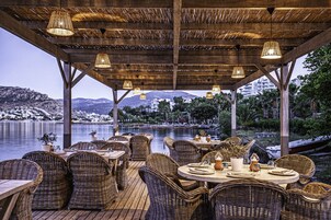 Un espacio de comedor al aire libre con mobiliario de mimbre y luces colgantes, con vistas a un cuerpo de agua y montañas.