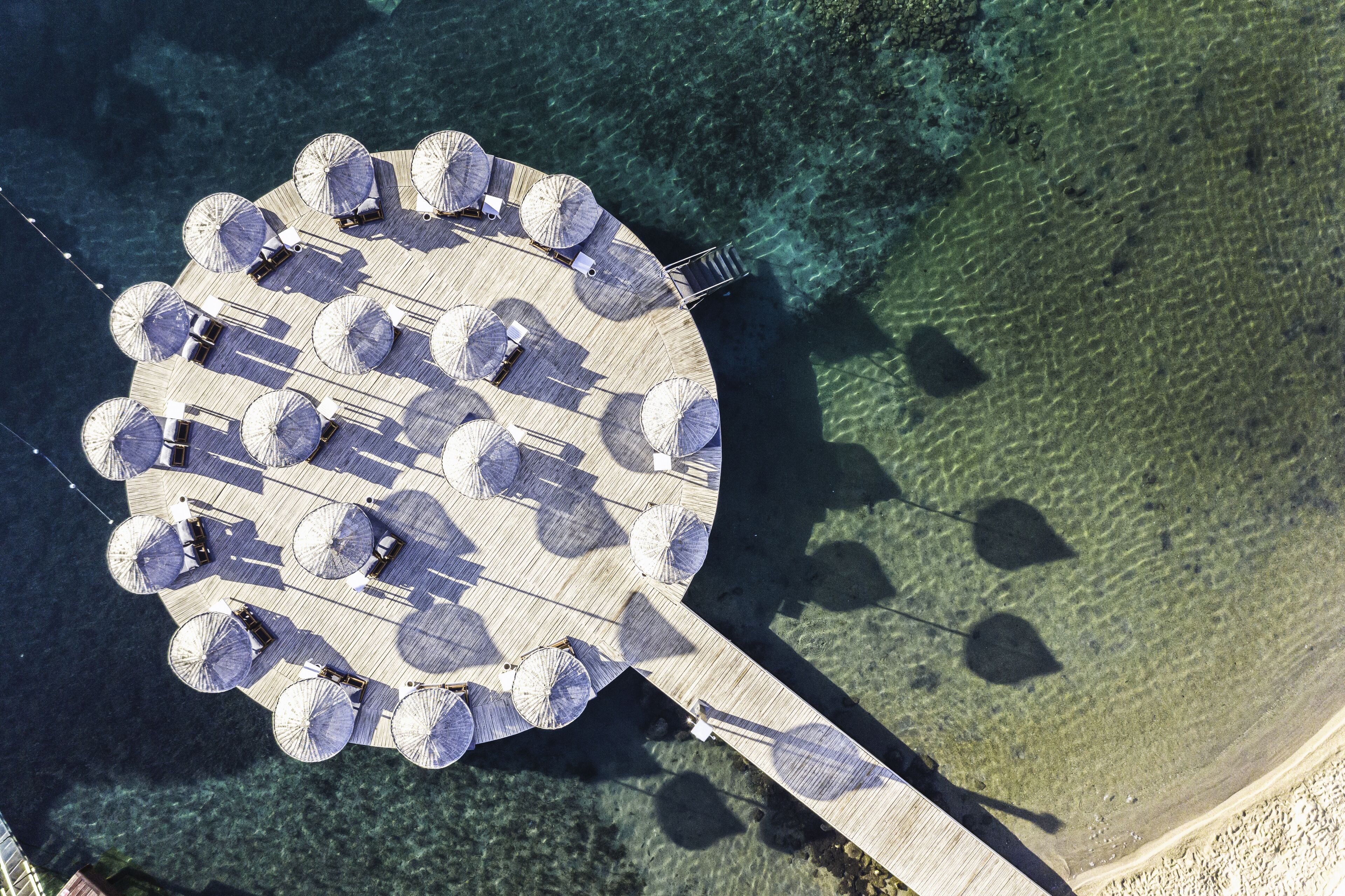 Vue aérienne d’une terrasse circulaire en bois, agrémentée de plusieurs parasols et de chaises longues, et entourée d’une eau cristalline.