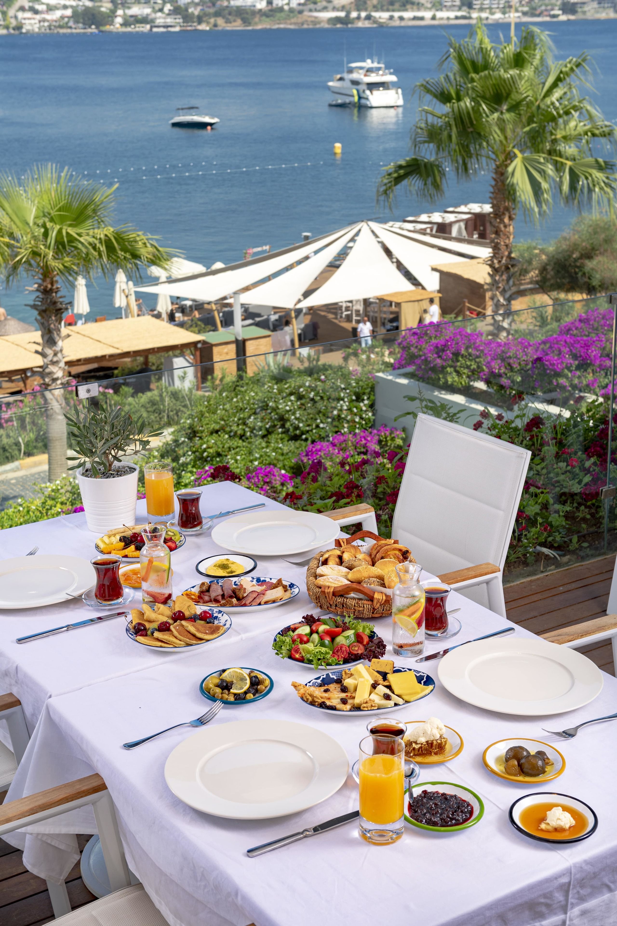 A table set for breakfast with various dishes, a glass of orange juice, and a view of the sea and boats.