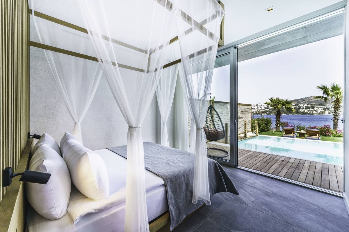 A bedroom with a bed, white curtains, and a view of a swimming pool and landscape.