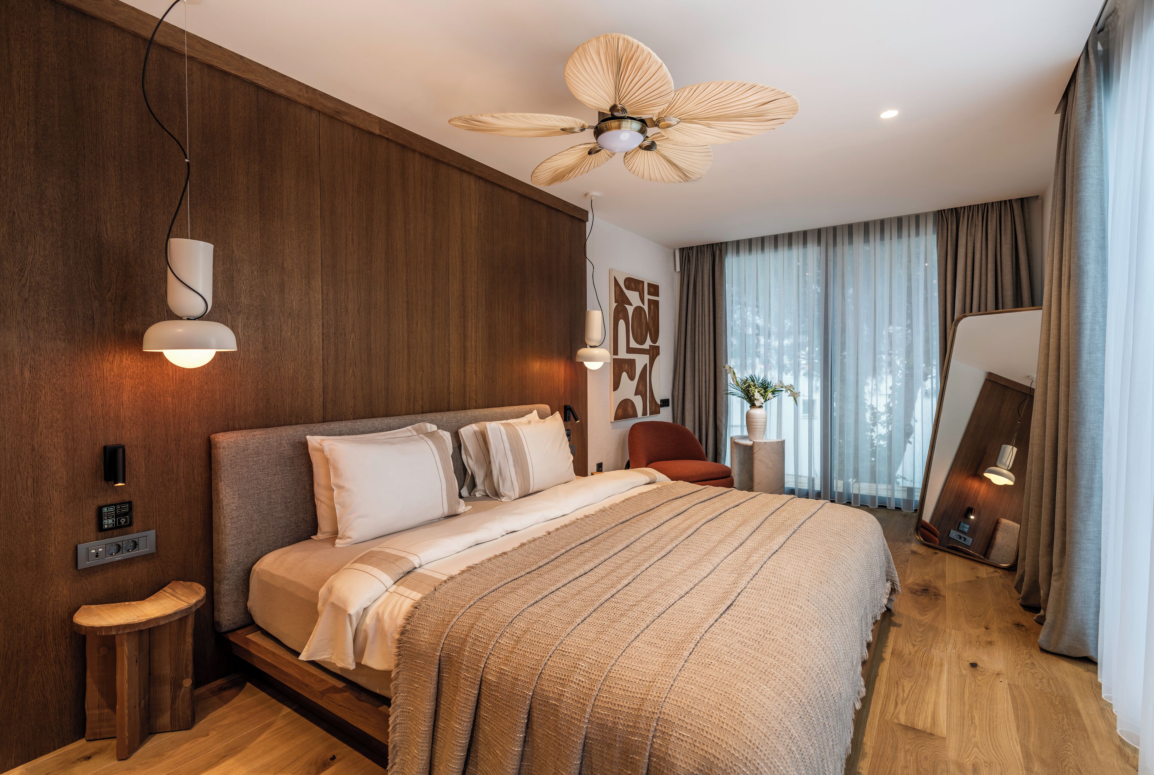 A modern bedroom with a large bed, wooden panel walls, a ceiling fan, and a bedside table with a lamp.