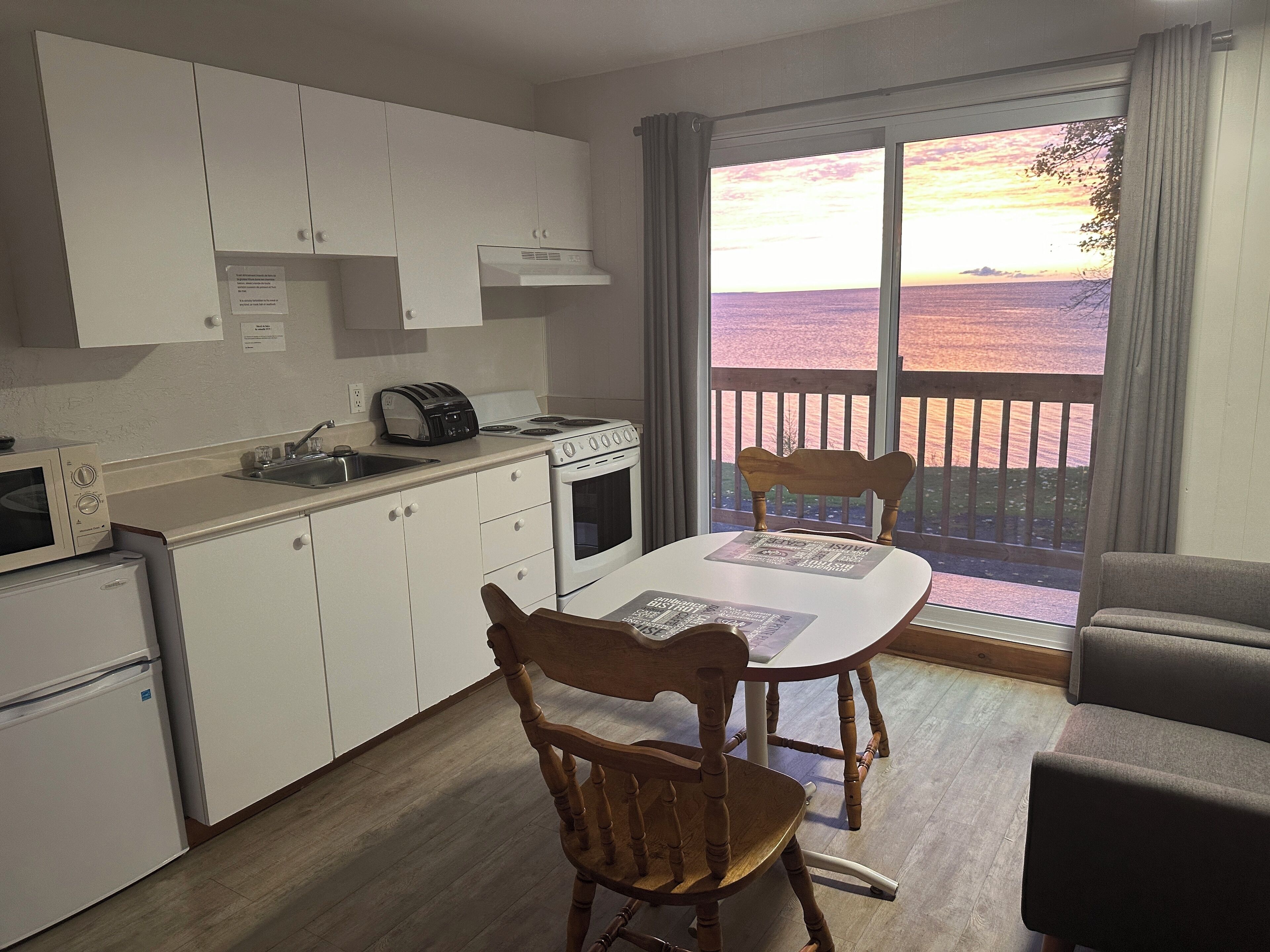 Une cuisine compacte avec des meubles blancs, une cuisinière et un grille-pain. On y trouve une table à manger avec deux chaises, et une grande fenêtre offre une vue sur la mer.
