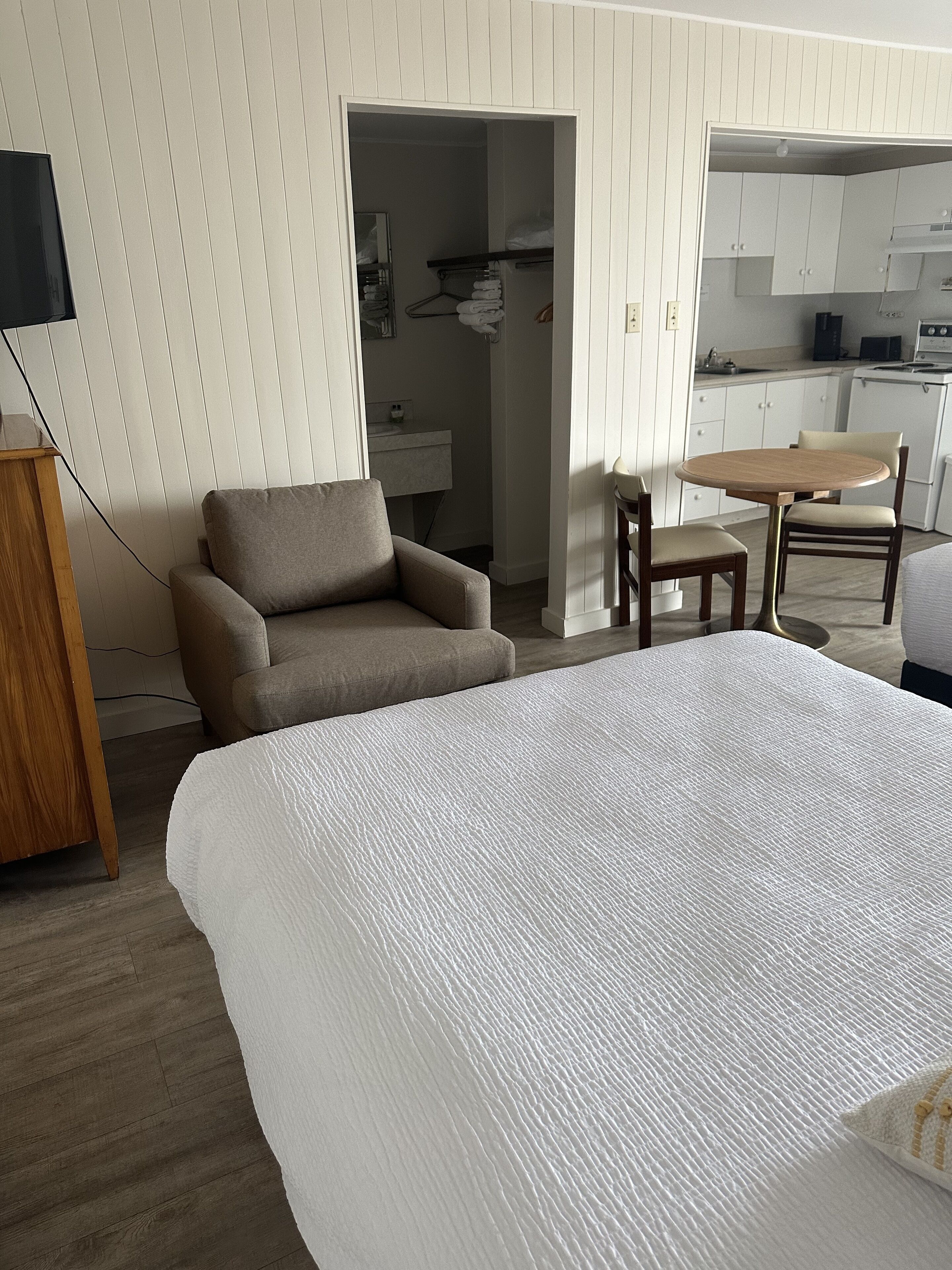 Een slaapkamer met een groot bed, een stoel, een tafeltje en een kitchenette op de achtergrond.