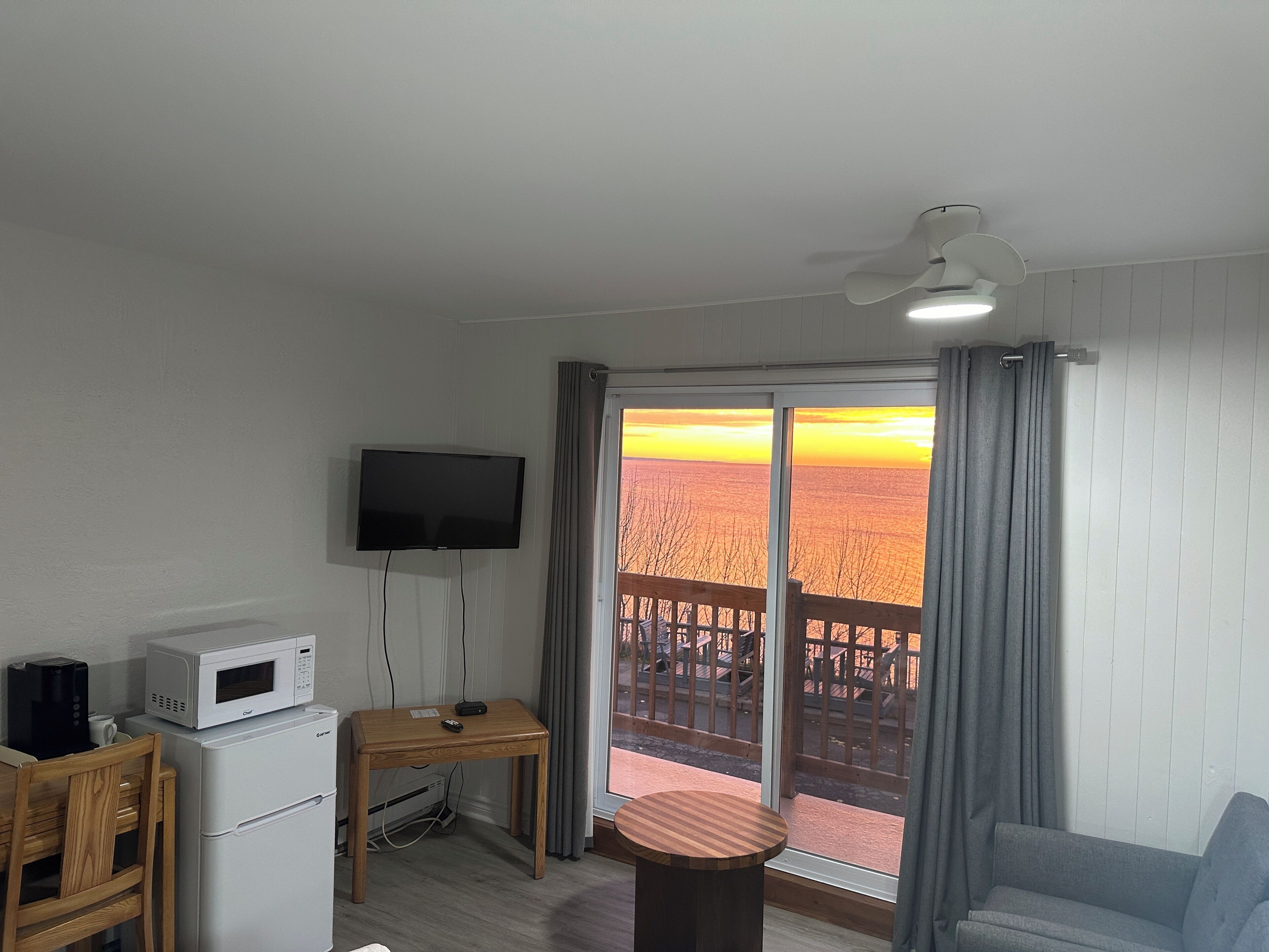 A living room with a flat-screen TV, a wooden table, a sofa, and a balcony with a view of a sunset over a forest.