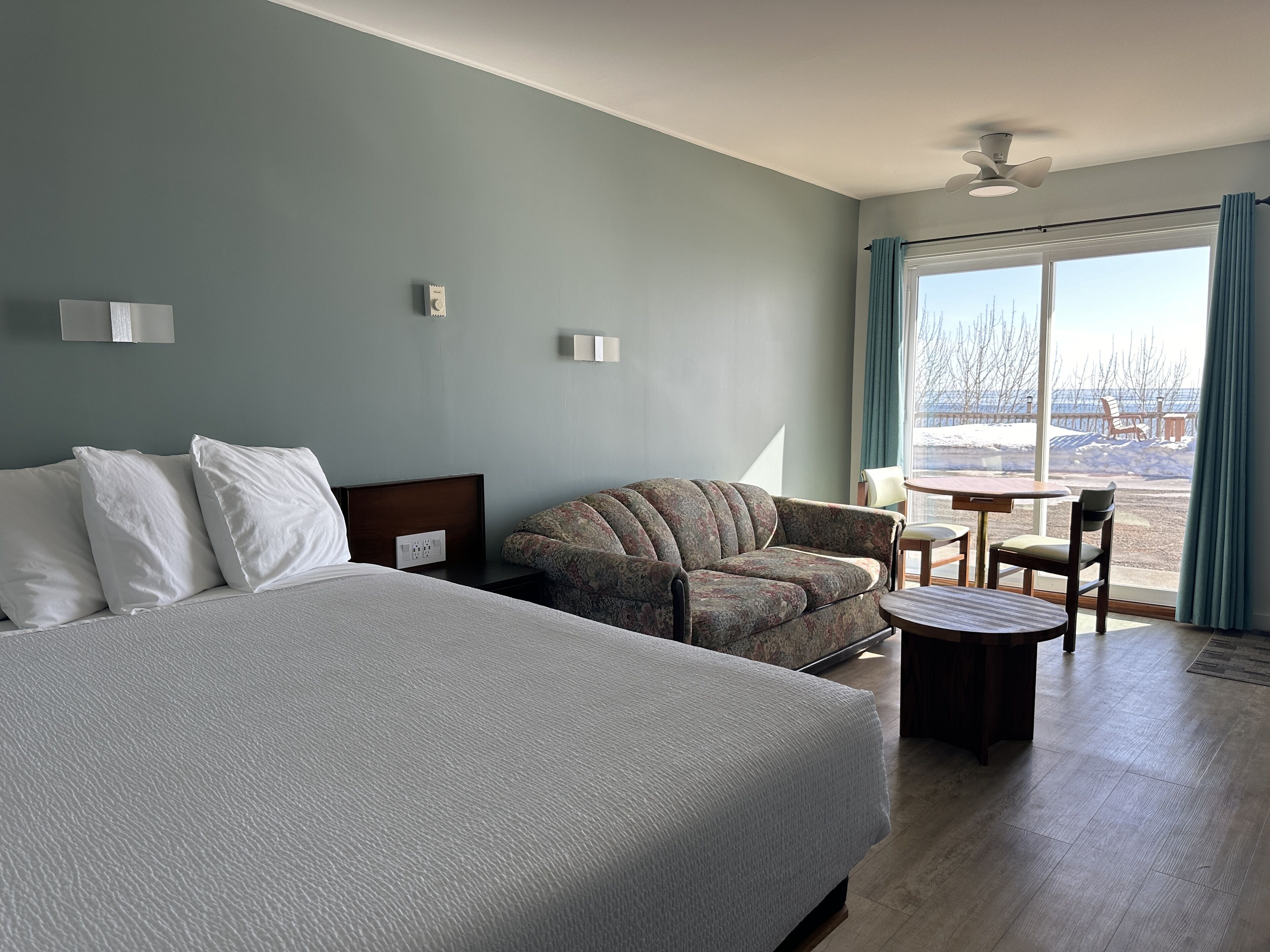 A hotel room with a large bed, a sofa, a small table, and a view of a snowy landscape through the window.