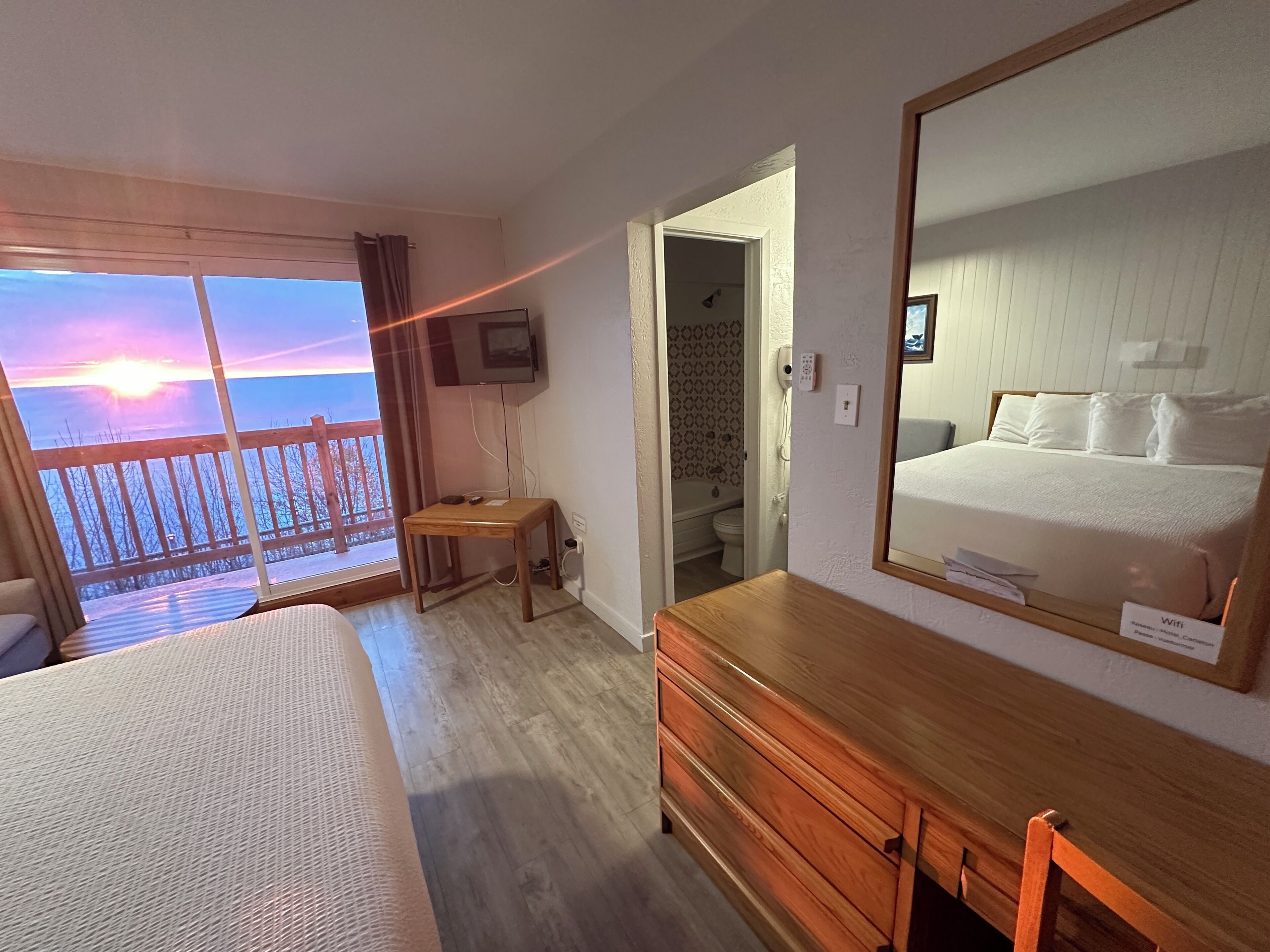 A hotel room with a bed, a wooden dresser, a television, and a balcony with a view of the sunset.