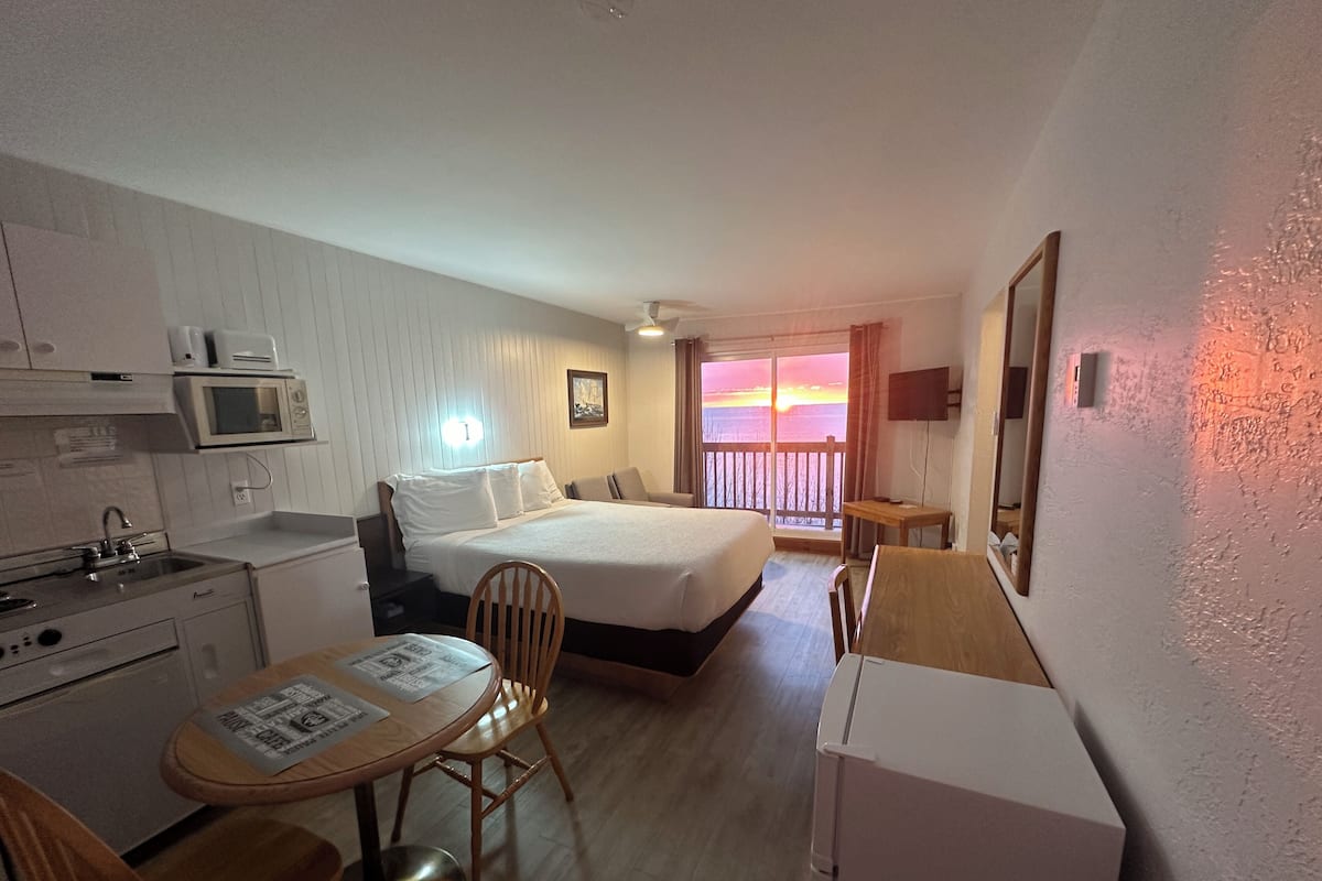 A hotel room with a bed, a desk, a chair, a microwave, a sink, and a balcony with a sunset view.