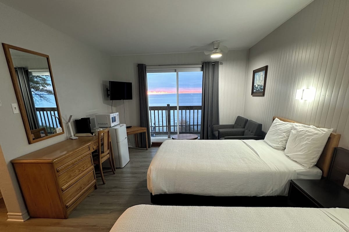 A hotel room with two beds, a desk, a TV, and a balcony with a view of the sea.