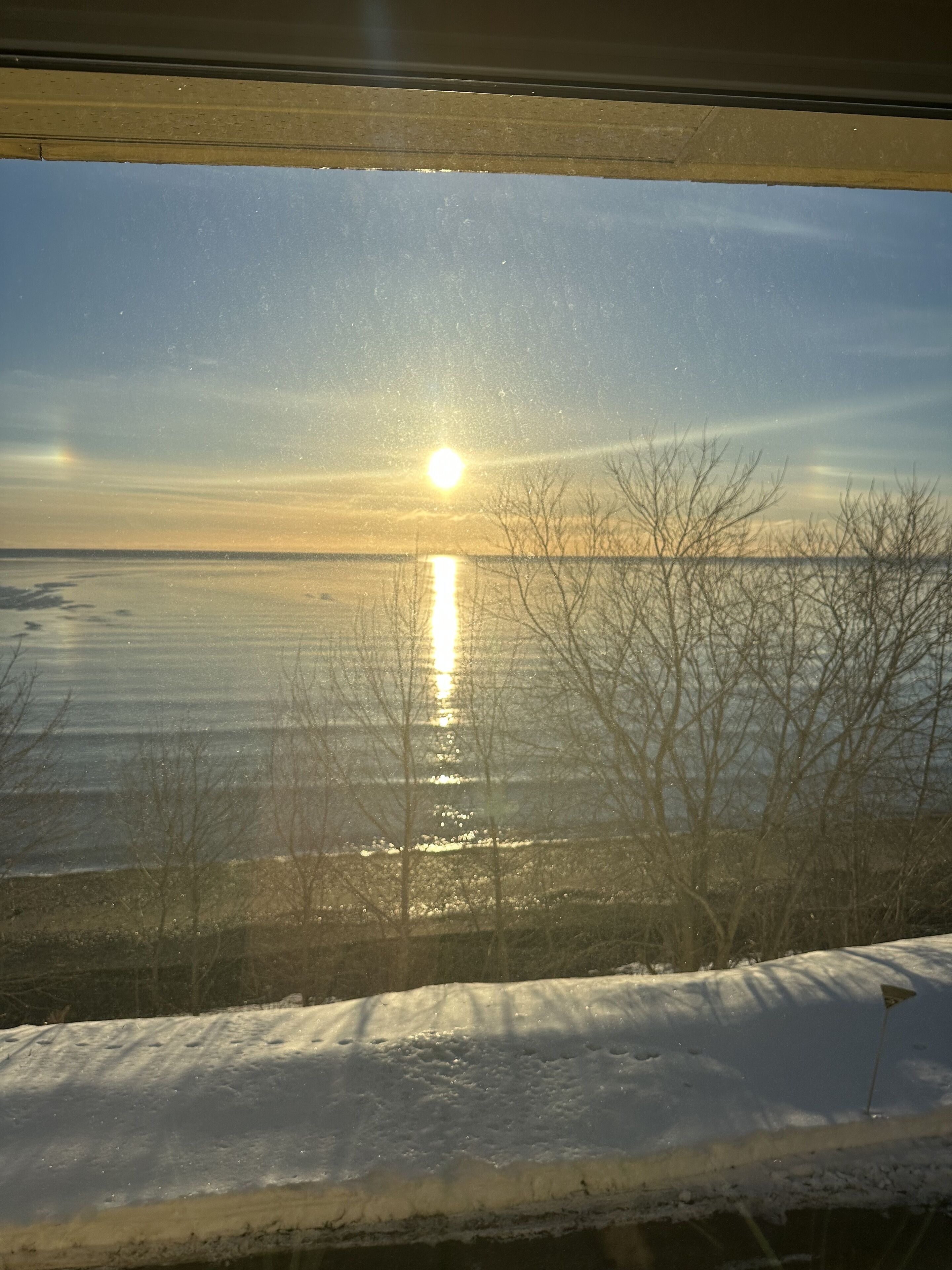Une vue à travers une fenêtre sur un paysage enneigé, avec des arbres sans feuilles et une étendue d’eau calme qui reflète le soleil.