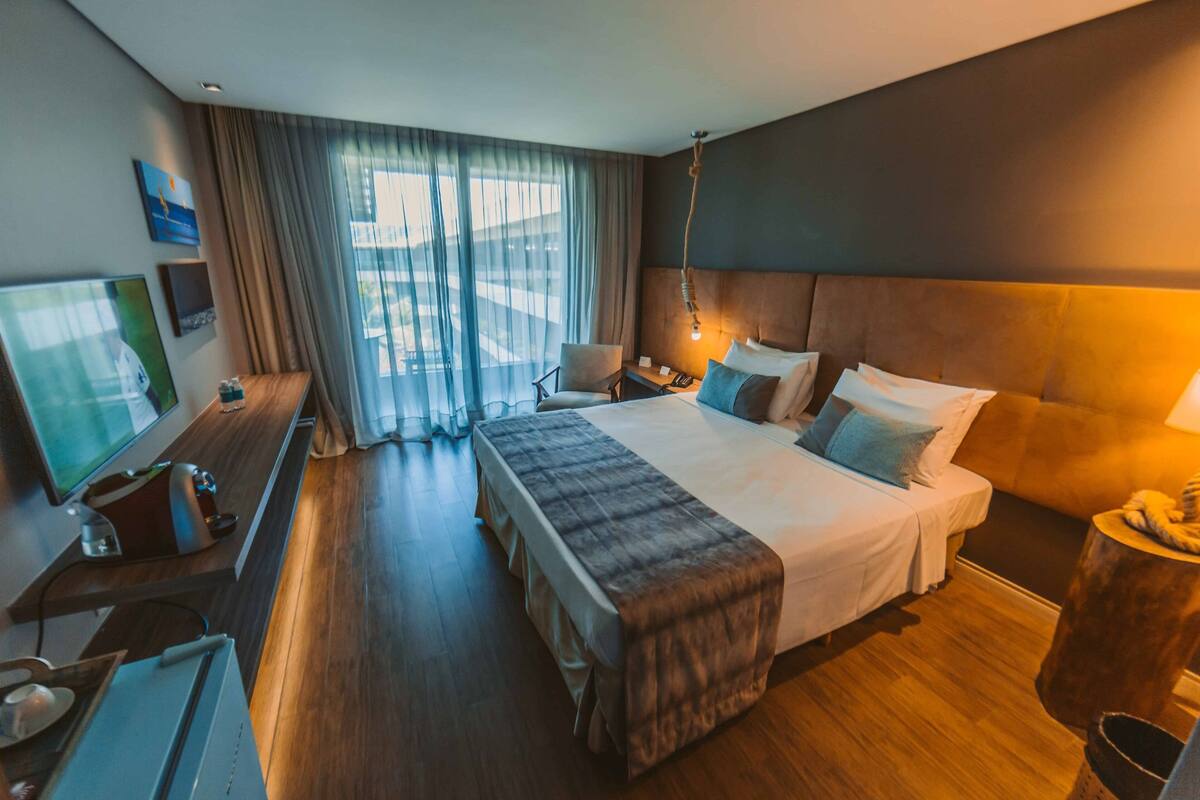 Habitación de hotel con una cama grande, un escritorio con computadora, televisión y ventana con cortinas.