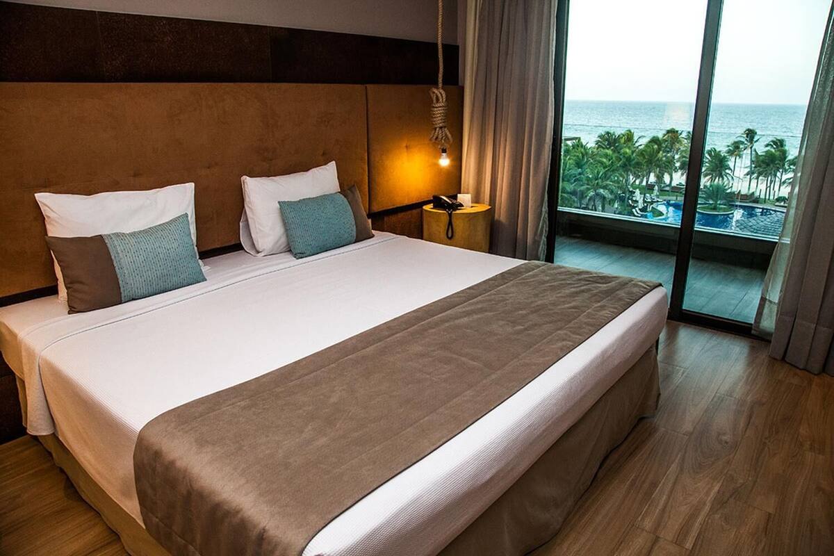 Una cama grande con cabecera marrón, almohadas blancas y azul turquesa, una lámpara de noche y vista al océano a través de ventanas amplias.