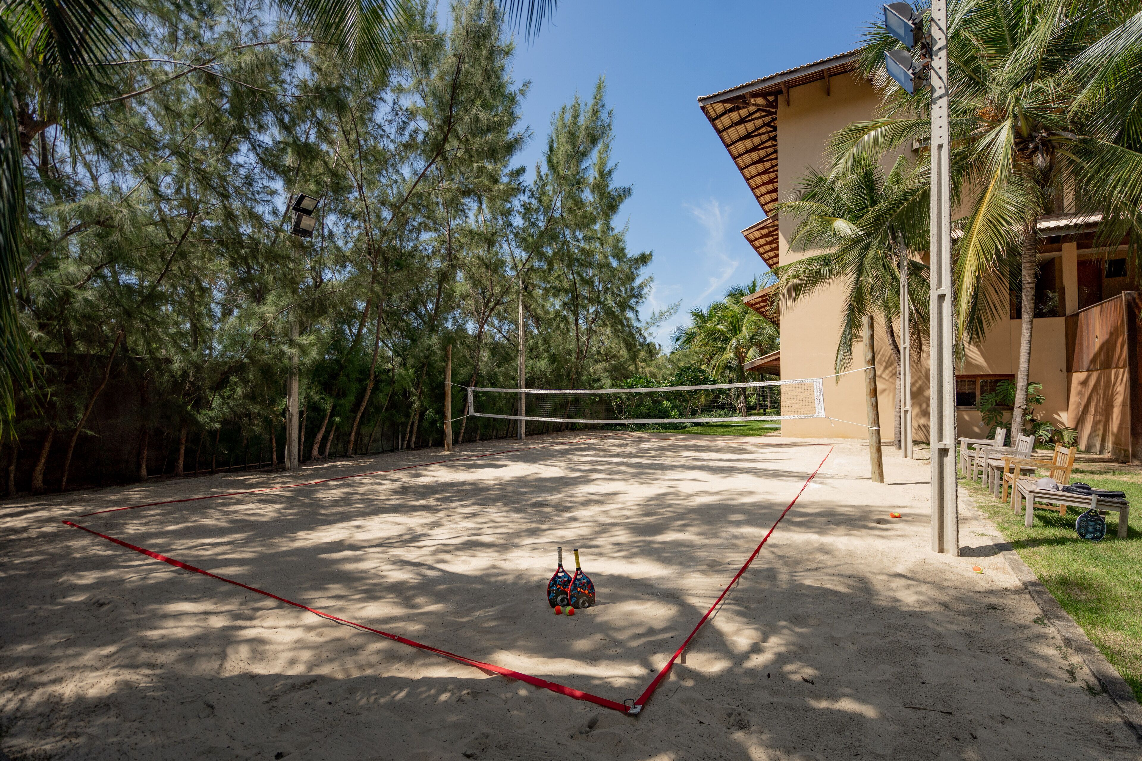A sandy volleyball court surrounded by tall trees and a building with a sloped roof.
