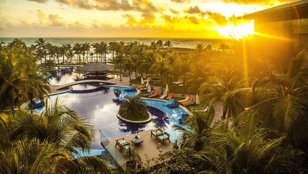 Un resort con una gran piscina, palmeras y vistas al océano al atardecer.