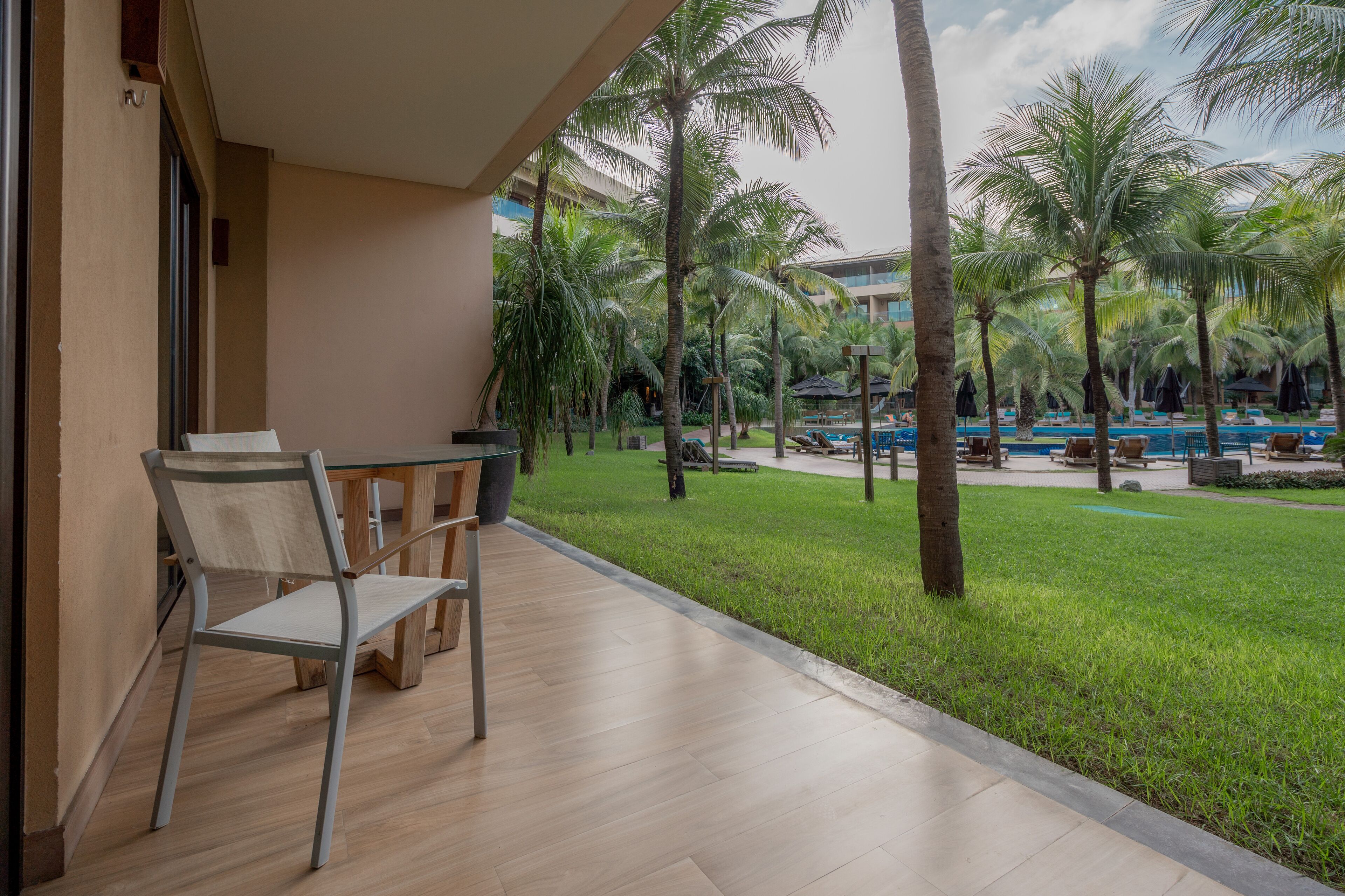 A balcony with a table and chairs, overlooking a pool area with palm trees and a grassy lawn.