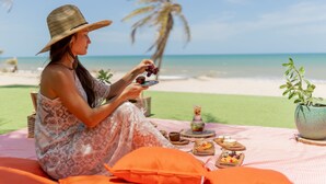 Una persona sentada en la playa con un montaje de picnic que incluye comida y bebidas, palmeras y el océano al fondo.