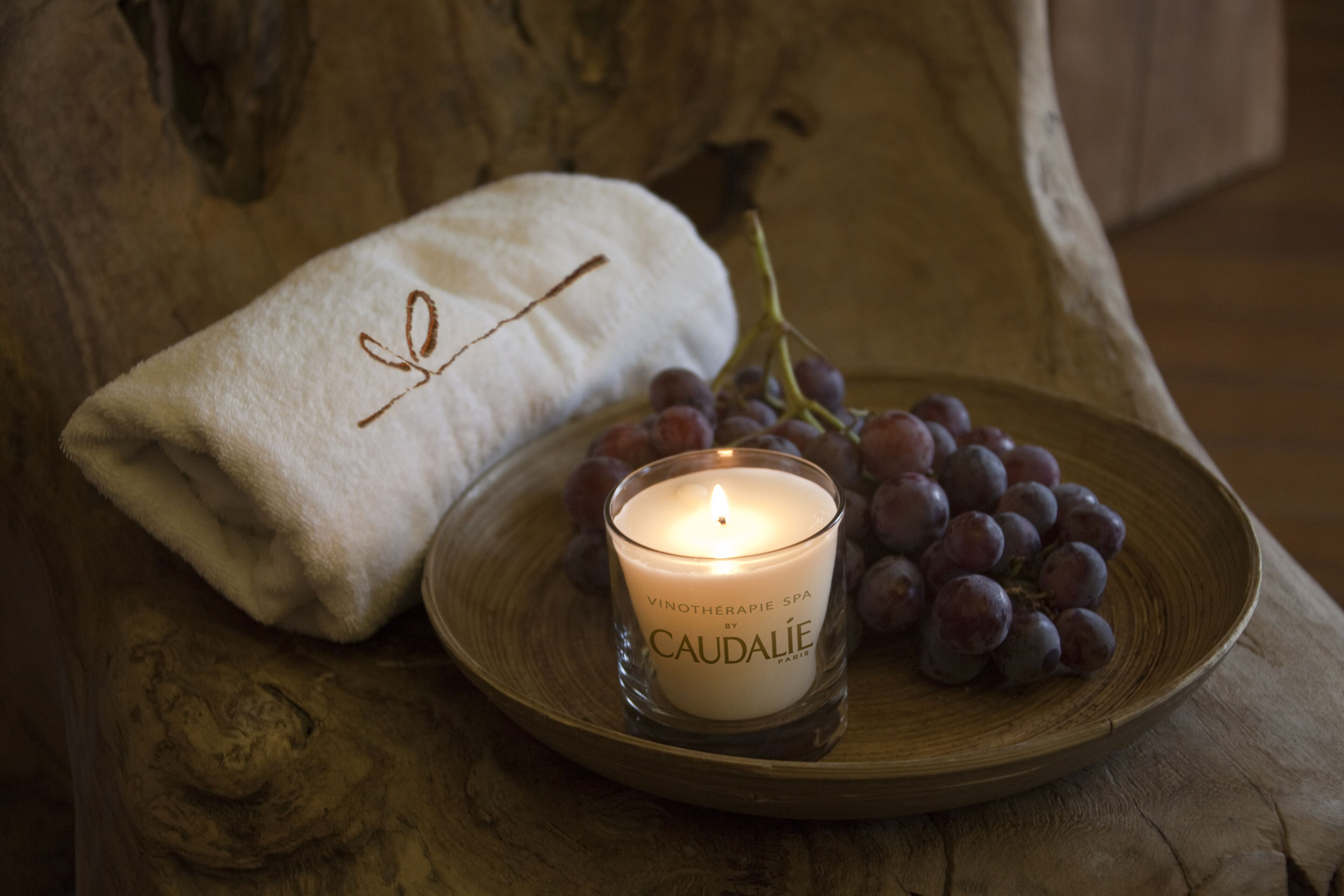 A candle labeled "Caudalie" on a wooden tray, a white towel with a leaf design, and a bunch of grapes.
