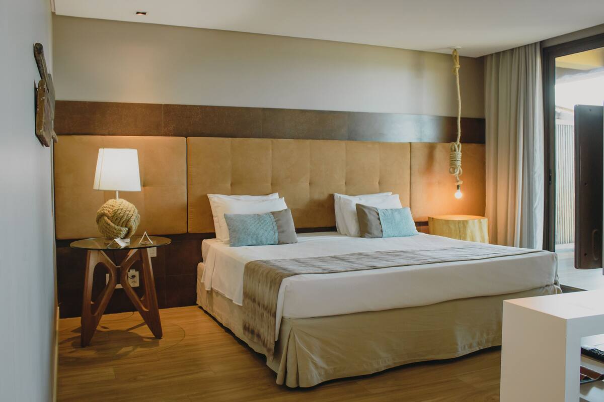 Habitación de hotel con una cama grande, una mesita de noche con lámpara, suelo de madera y vistas al exterior.