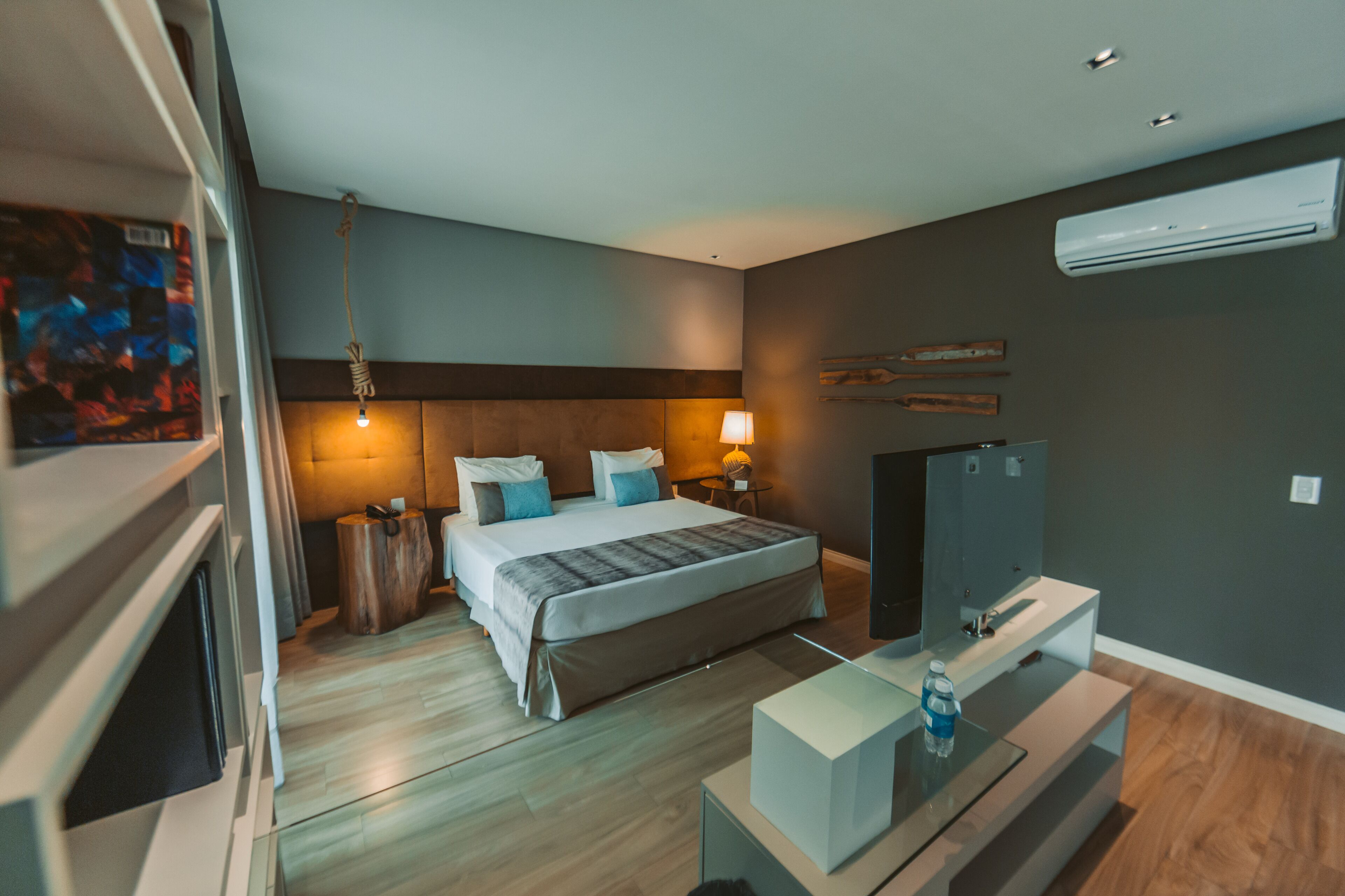 A modern hotel room with a large bed, bedside lamps, a flat-screen TV, a small table with a water bottle, and an air conditioning unit on the wall.