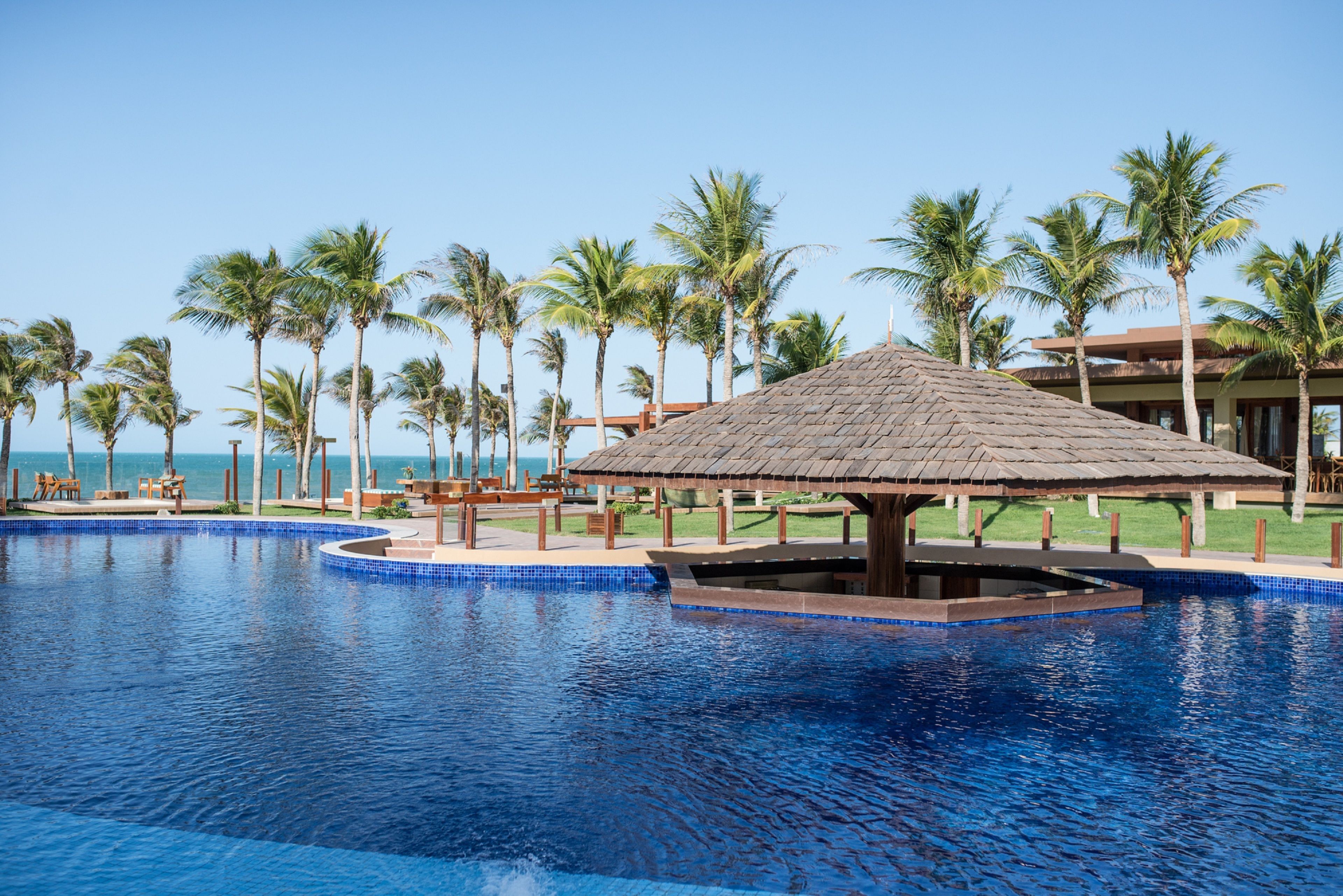 A pool with a wooden gazebo, palm trees, and a view of the ocean.