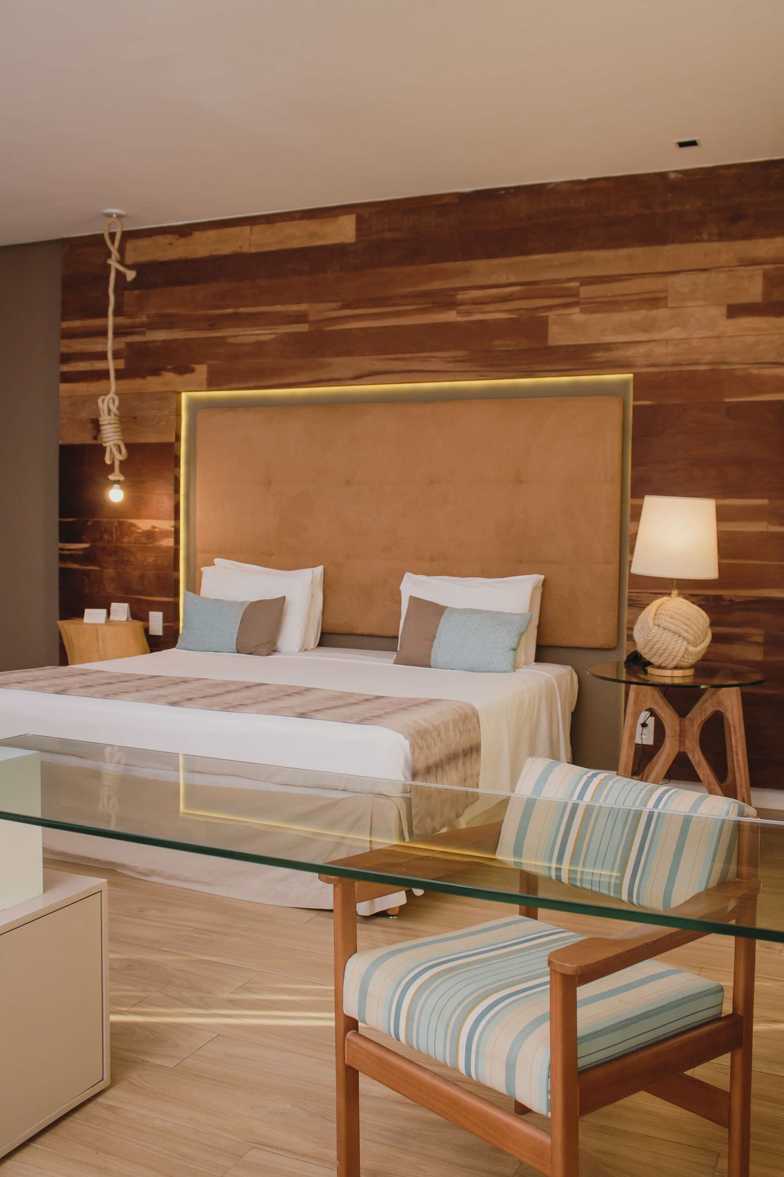 A modern hotel room with a large bed, a glass table, a wooden chair, and a wooden headboard.
