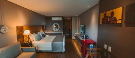 Habitación de hotel con cama, sofá, escritorio con silla, televisor y obras de arte en las paredes.