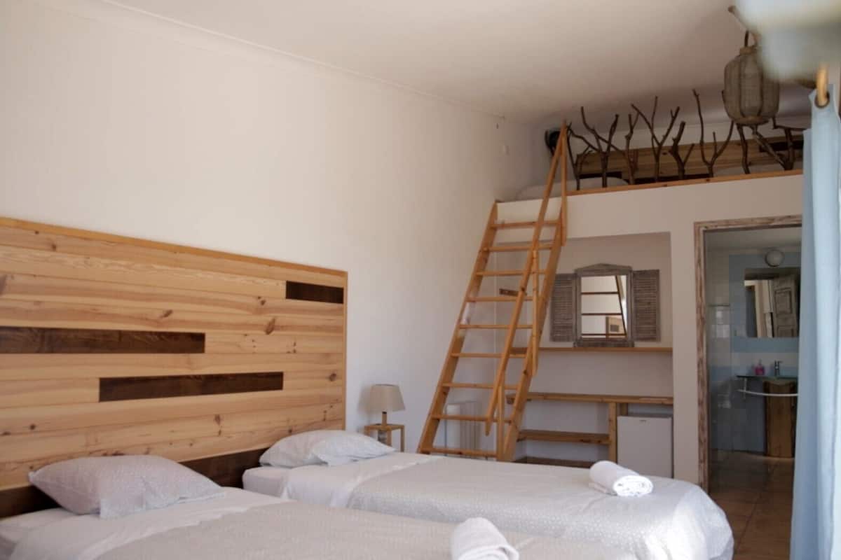 Une chambre avec une tête de lit en bois, deux lits avec du linge de lit blanc, une échelle en bois, une petite table avec une lampe, et une vue sur une salle de bain par une porte ouverte.