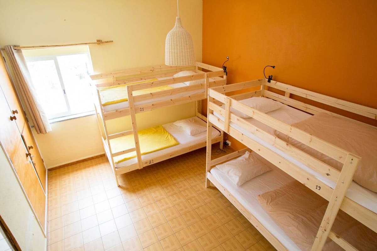 Une pièce avec trois lits superposés, un plancher en bois, une fenêtre avec des rideaux et un luminaire suspendu.