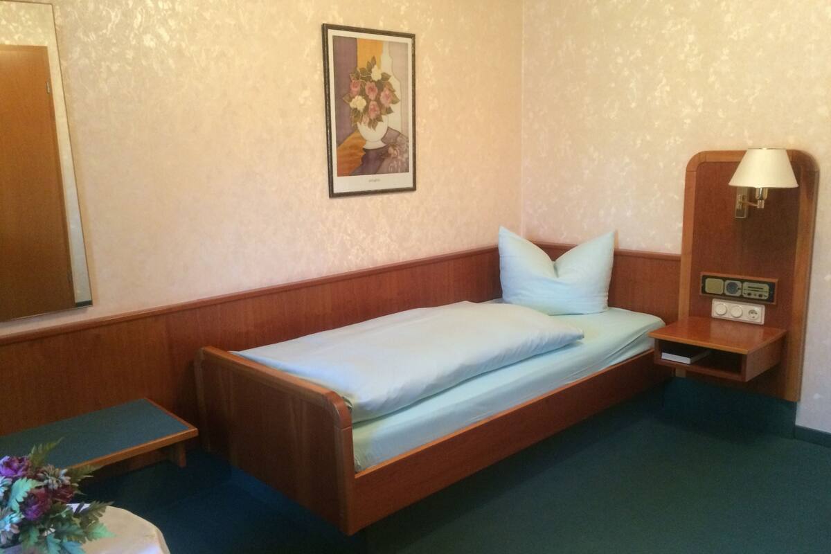 A single bed with a wooden headboard, a bedside table with a radio, and a small table with a vase of flowers.