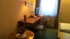 A hotel room with a wooden desk, a chair, a small table with a vase of flowers, a lamp, a television, and a bed. - Gästehaus Wild (Oberasbach)