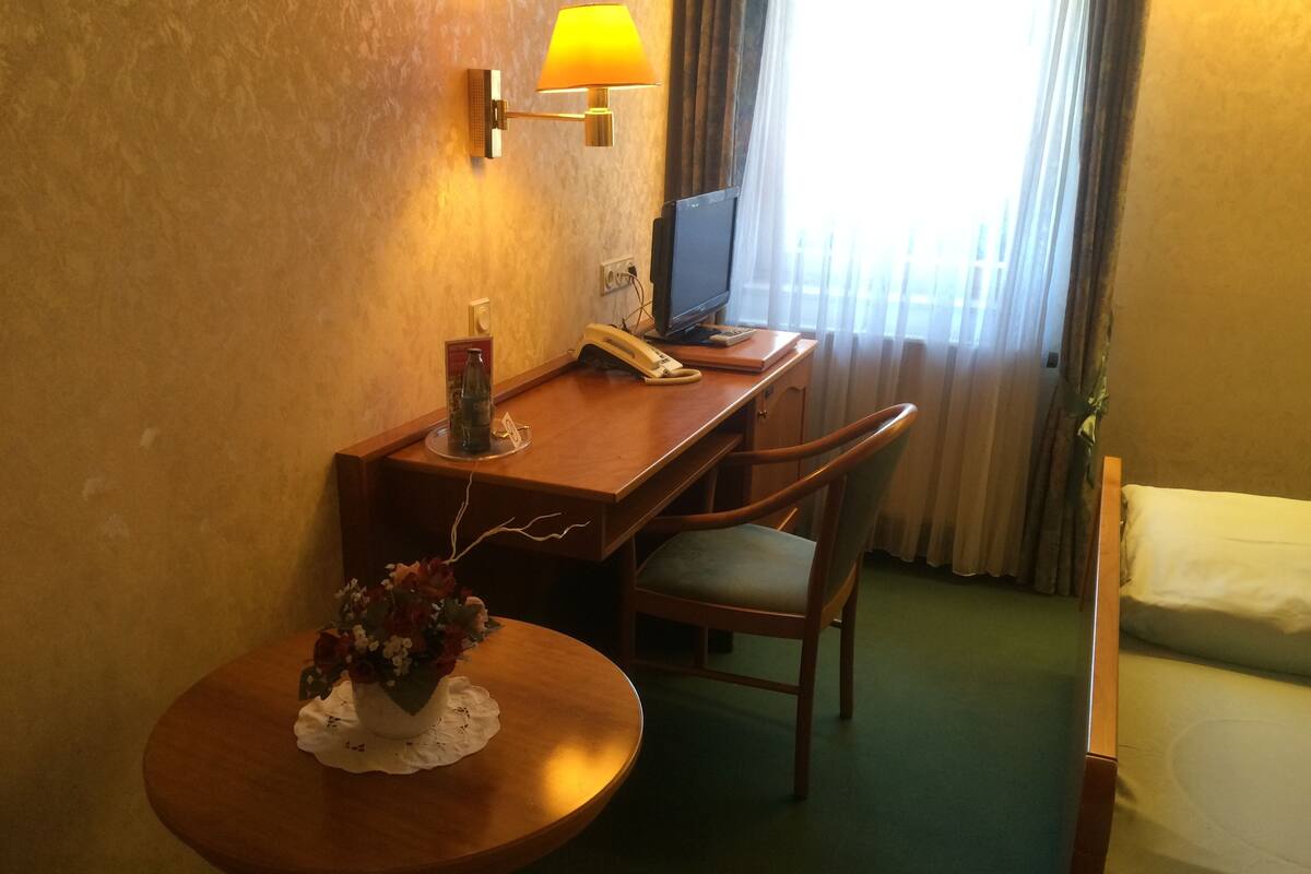 A hotel room with a wooden desk, a chair, a small table with a vase of flowers, a lamp, a television, and a bed.