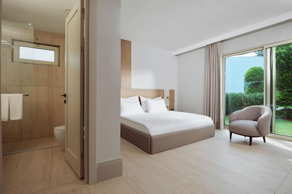 Une chambre d’hôtel moderne dotée d’un grand lit, d’une salle de bain avec toilettes et douche, et offrant une vue sur l’extérieur grâce à une grande fenêtre.