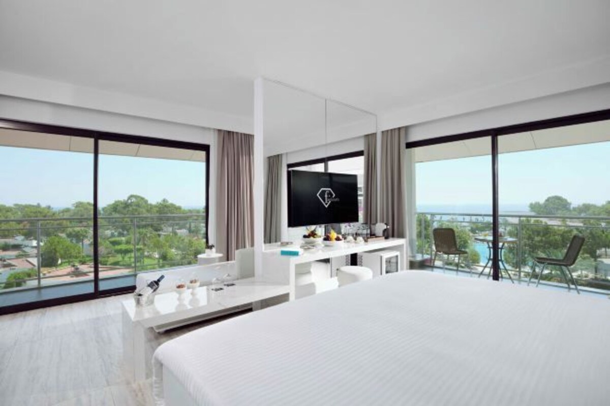 A modern hotel room with a large bed, a flat-screen TV, a desk with a chair, and a balcony with a view of the city.