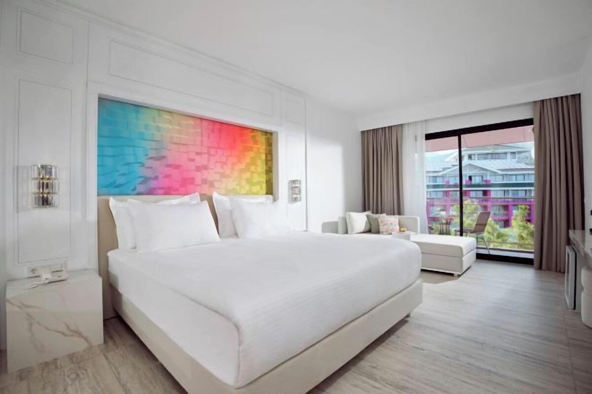 Une chambre d’hôtel moderne avec un grand lit, un canapé, une œuvre d’art colorée au mur et un balcon avec vue.