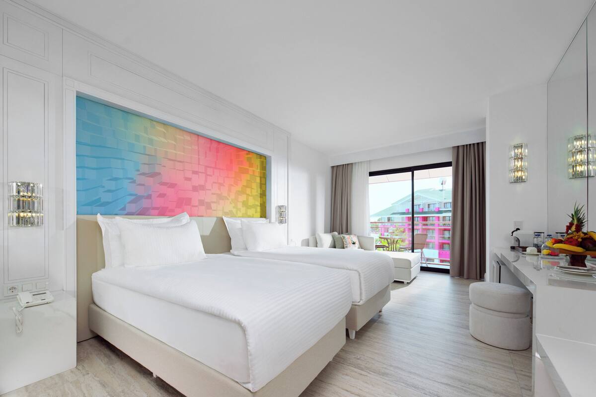 Une chambre d’hôtel moderne avec deux lits, une grande fenêtre offrant une belle vue, un bureau avec une chaise et une œuvre d’art colorée accrochée au mur.