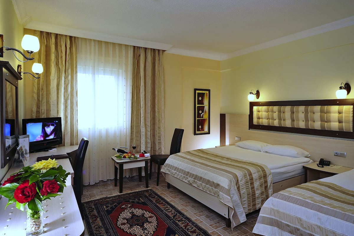 A hotel room with a bed, a desk with a computer, a chair, a small table with fruits and a glass of wine, and a decorative vase with flowers.