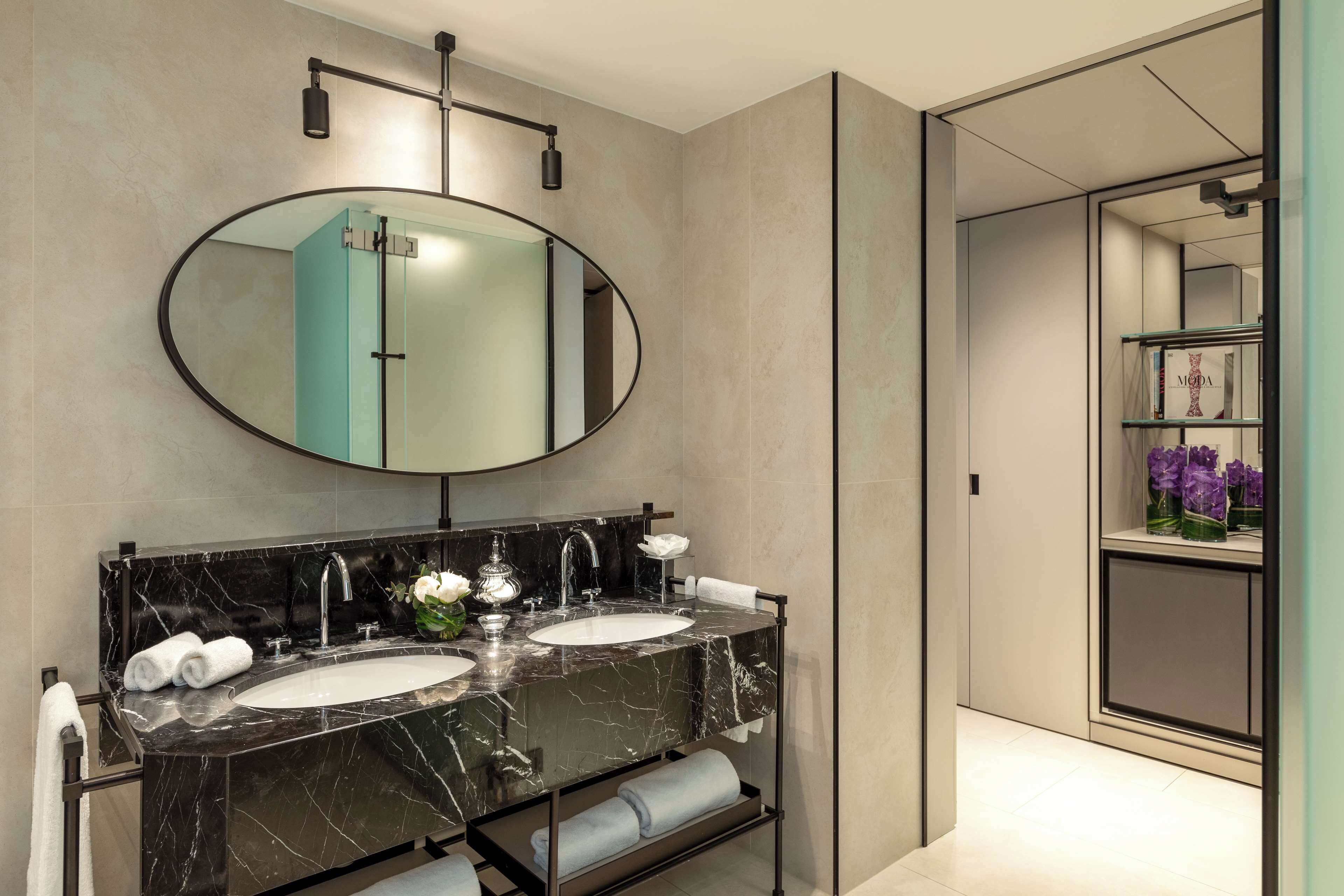 A modern bathroom with a large oval mirror, two sinks, and a vanity with a marble countertop.