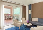 A modern hotel room with a balcony, a sofa, two armchairs, a small round table, and a vase of flowers.