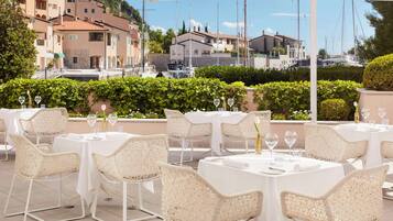 Tavoli da pranzo all'aperto con sedie in vimini, vista sul porto turistico e edifici sullo sfondo.