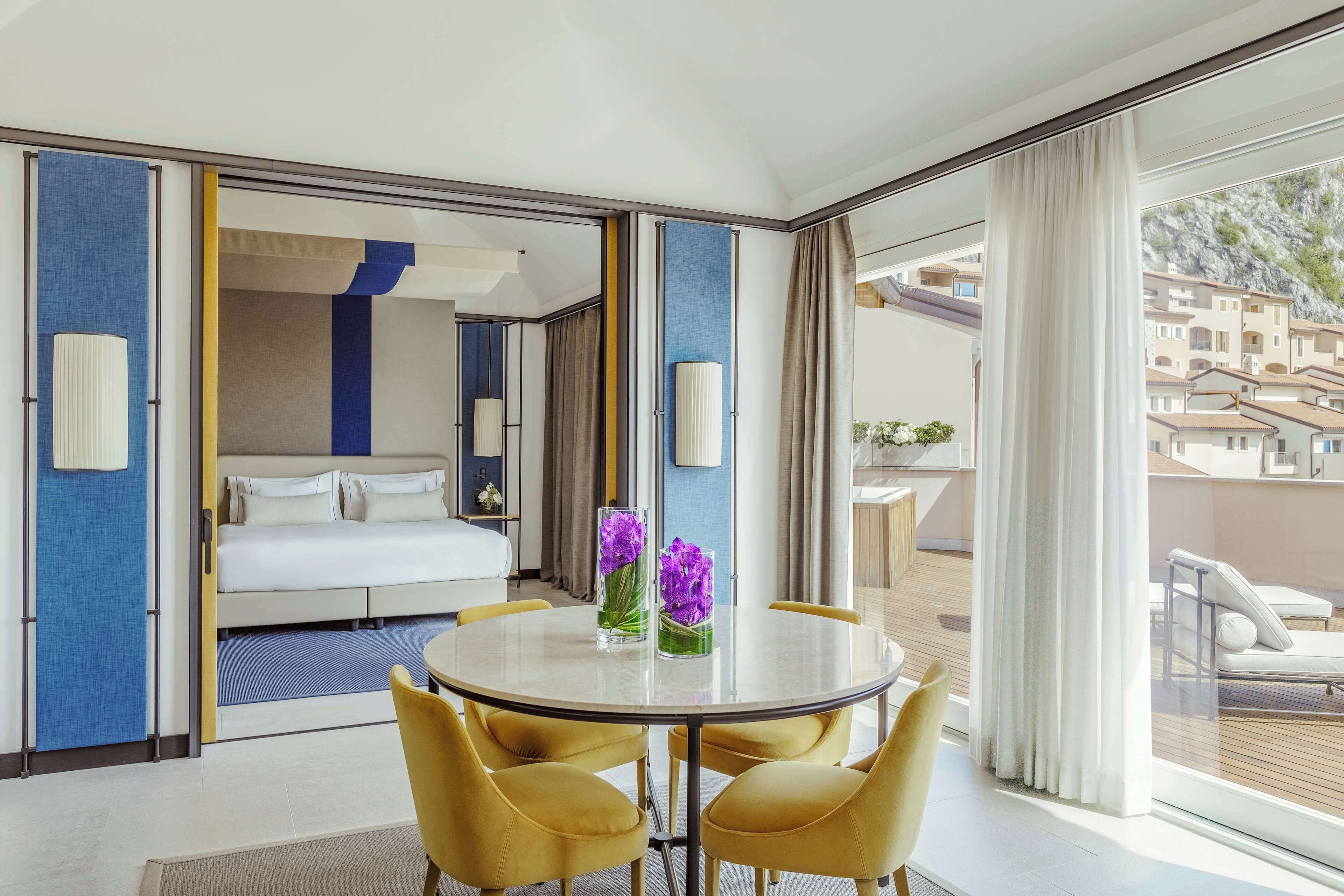 A hotel room with a bed, a round table with chairs, and a balcony with a view of buildings.