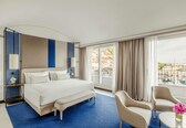 A hotel room with a large bed, two armchairs, a small table, and a view of buildings and water through the window.